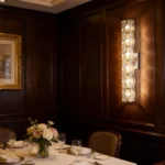 Illuminated crystal wall sconce mounted on dark wood paneling beside a formal dining table