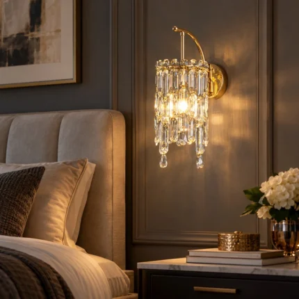 Gold crystal wall sconce Style A with hanging crystal prisms, installed beside a bed in a warm bedroom setting